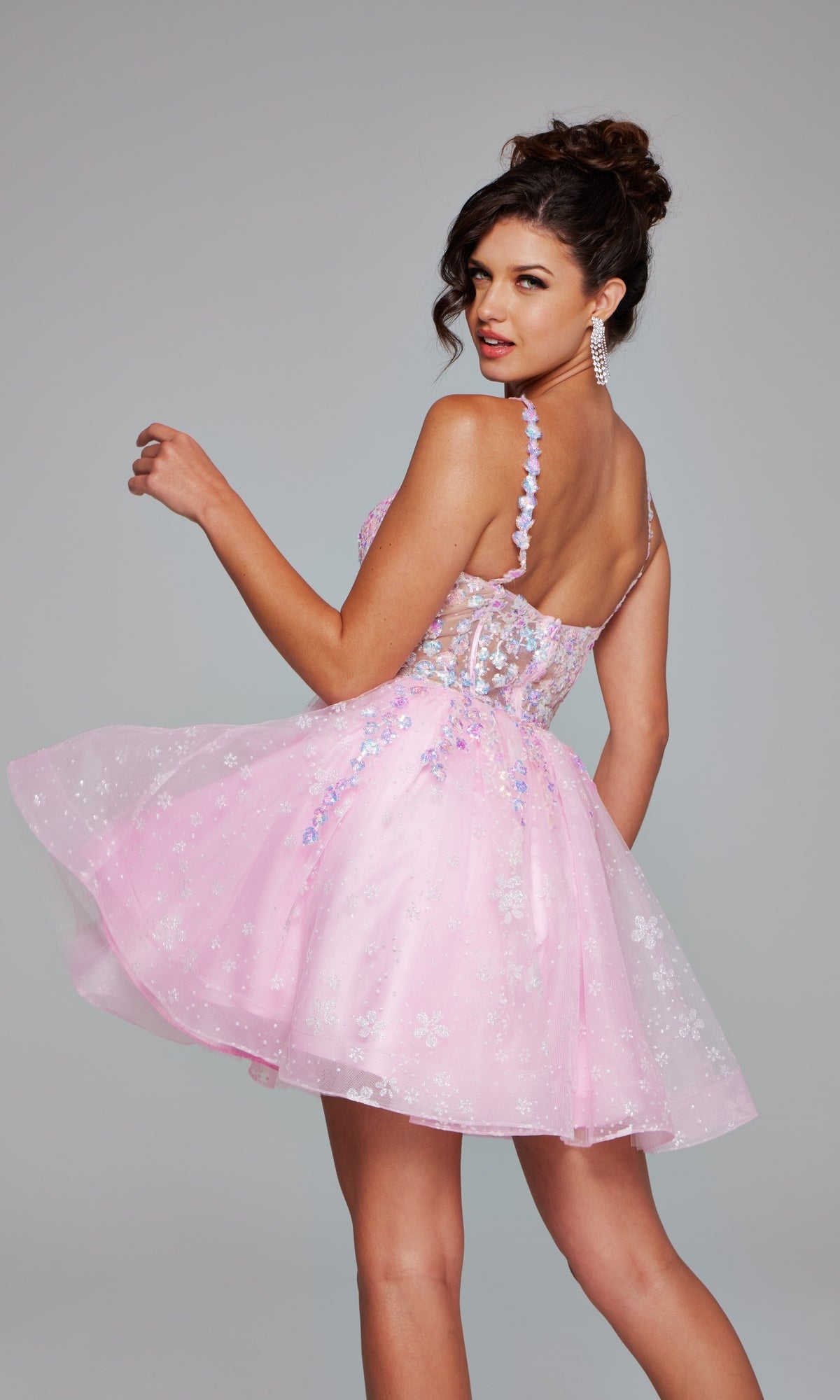 A woman poses in the Short Homecoming Dress 40501 by Jovani, featuring pink floral sequin embroidery and sheer accents, looking over her shoulder against a plain background.